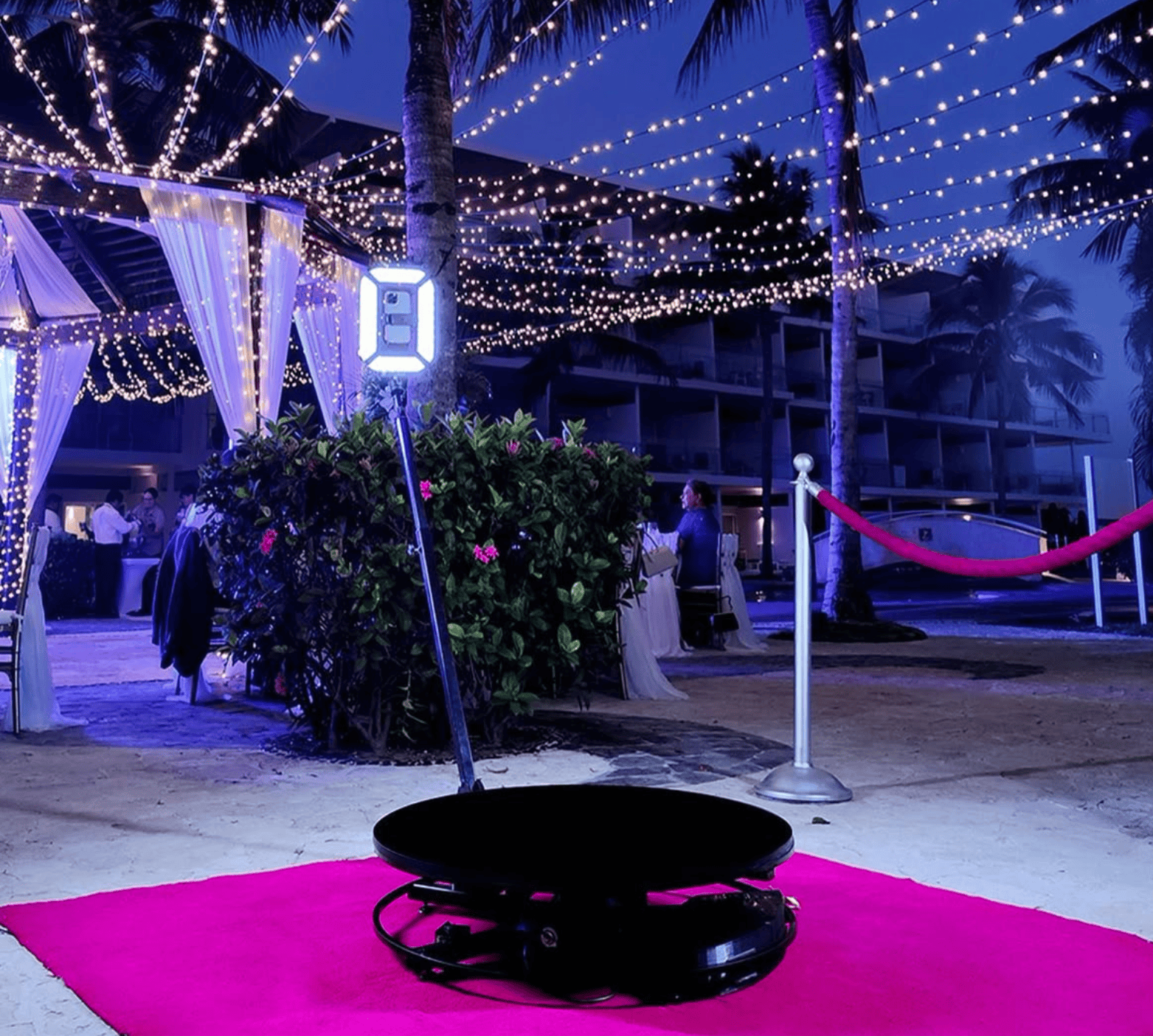 360 photo booth platform on a pink carpet, with its illuminated arm and a rope stanchion, at an outdoor evening reception under string lights and palm trees