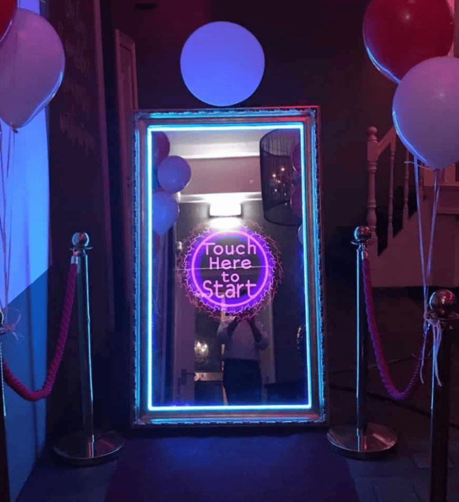Magic mirror photo booth with a glowing blue frame displaying “Touch Here to Start”, flanked by two stanchions with red ropes and balloons, in a dimly lit reception room