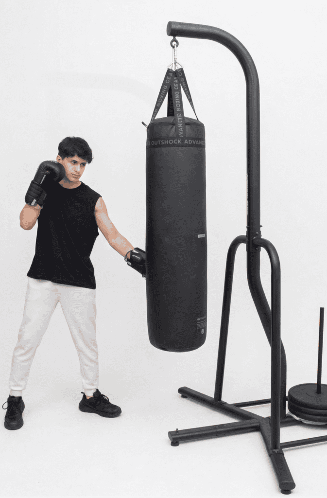 Young man in boxing gloves hitting a black punching bag hung from a freestanding stand weighted with plates, on a white background