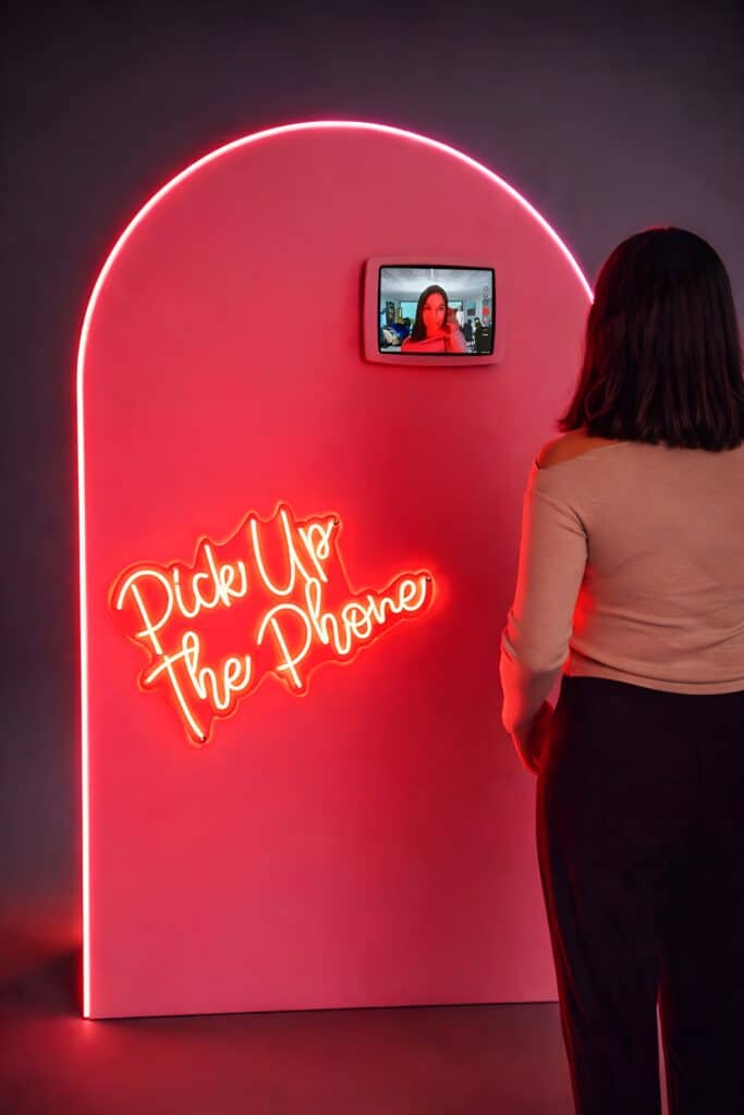 Attendee standing at a pink illuminated arch-shaped audio and video guest book with a tablet and a “Pick Up The Phone” neon sign