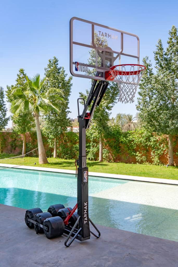 Mobile NBA basketball hoop with a weighted wheeled base, set up beside a swimming pool in a leafy garden