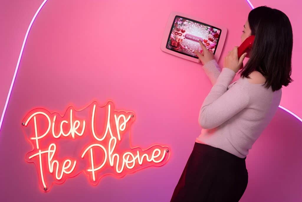 A guest holds a red handset to her ear and taps a tablet screen to record a message on an audio and video guest book, beside a “Pick Up The Phone” neon sign