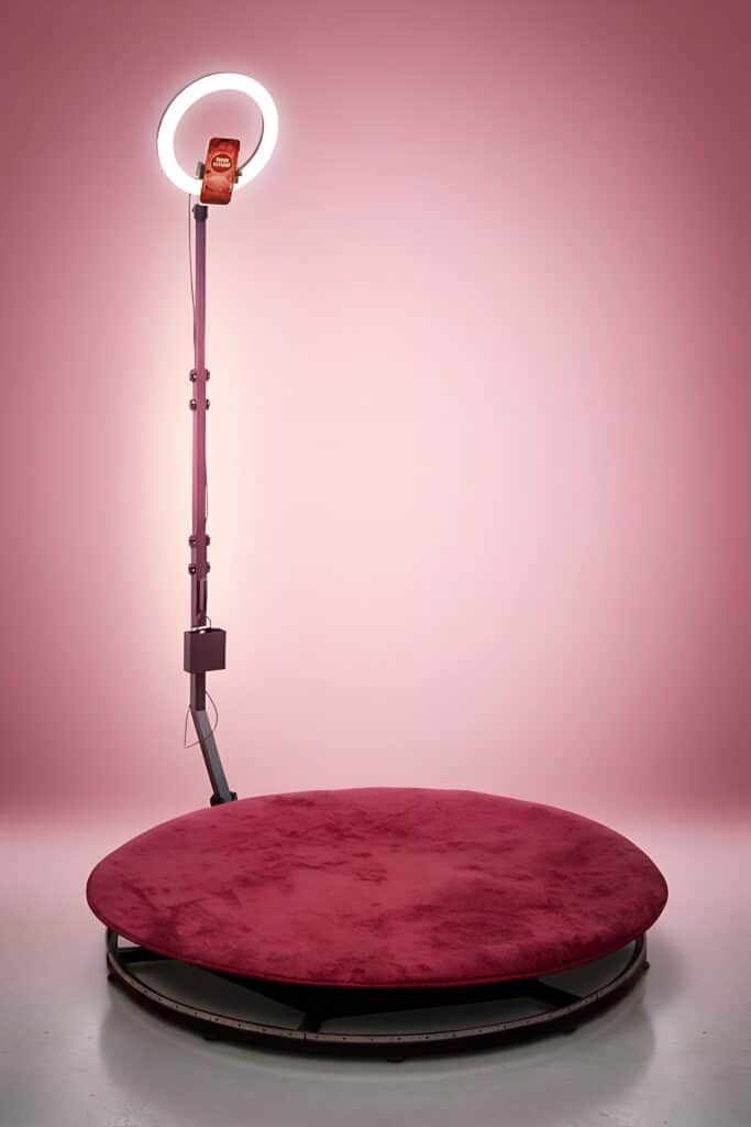 360 photobooth: round platform covered in red velvet and a pole with a ring light and smartphone, against a pink background